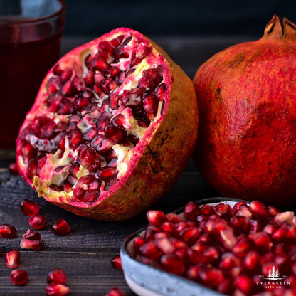 A pomegranate tree seedling with a half pomegranate fruit cut open to reveal the seeds inside, placed on a wooden surface with a whole pomegranate fruit and scattered seeds in the background.